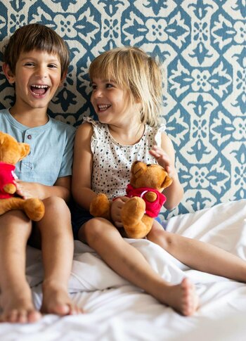 Zwei Kinder sitzen lachend auf dem Bett in einem hellen Hotelzimmer und halten Plüschtiere in den Händen.