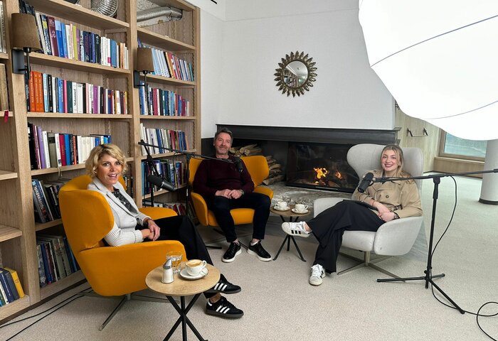 Three people talk in a cozy lounge with bookshelves and a fireplace, with microphones set up for the recording.