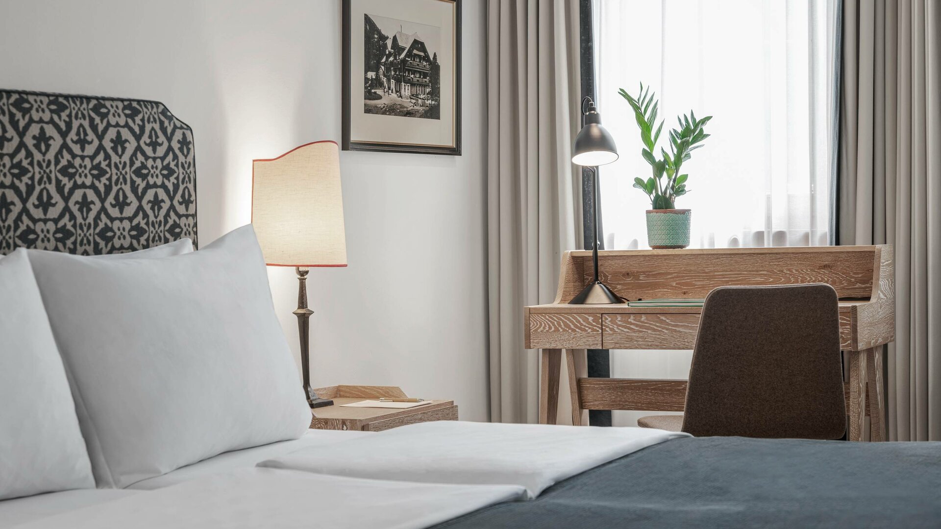 Ein Doppelbett mit gemachtem Bettzeug, Schreibtisch, Stuhl und Lampe in einem hellen Hotelzimmer.