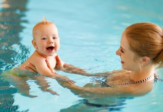 Eine Frau hält ein lächelndes Baby im Wasser eines Innenpools.
