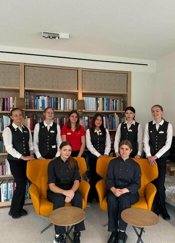 Praktikumsbetrieb im Hotel Hochschober Acht junge Frauen in Service- und Arbeitskleidung stehen und sitzen in einem Aufenthaltsraum mit Bücherregalen und Sesseln.