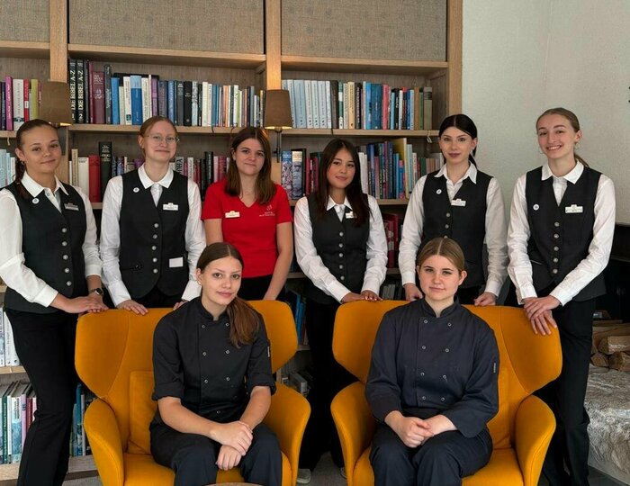 Praktikumsbetrieb im Hotel Hochschober Acht junge Frauen in Service- und Arbeitskleidung stehen und sitzen in einem Aufenthaltsraum mit Bücherregalen und Sesseln.