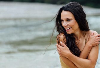 A smiling woman in a bikini sits by the water and holds herself with crossed arms, with calm water behind her.