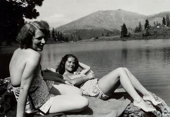 Two women sit relaxed on a rock by the lakeshore with mountains in the background.