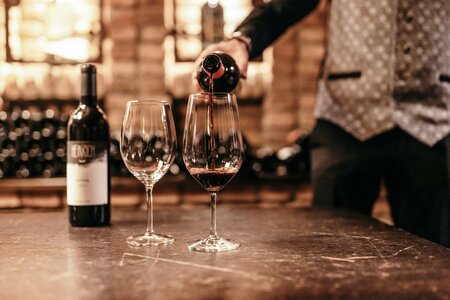 Eine Person schenkt Rotwein in zwei Gläser an einem Tisch ein, daneben steht eine Weinflasche.