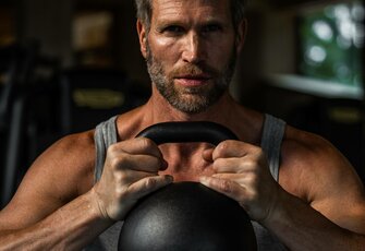 Ein Mann hält im Fitnessbereich eine Kettlebell mit beiden Händen vor der Brust.