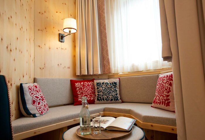 Gemütliche Sitzecke im Deluxe Zirbenzimmer mit Eckbank, Kissen, Holzwand, Fenster und kleinem Tisch mit Buch und Glasflasche.