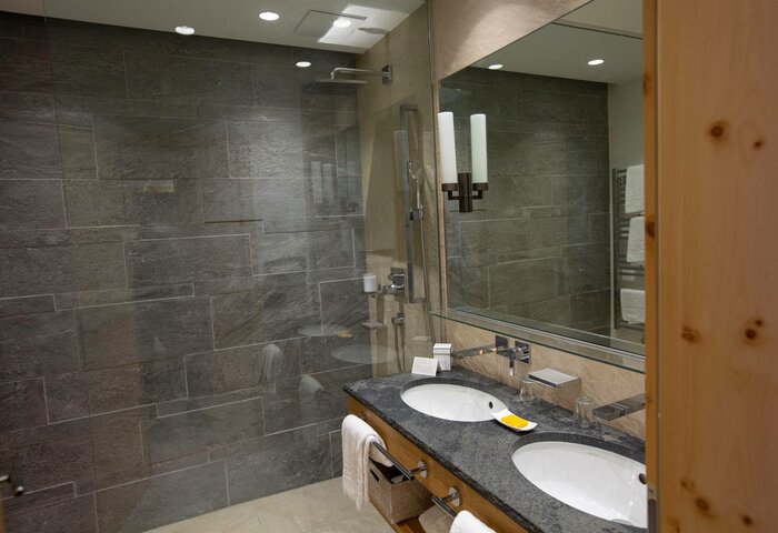 Das Bad im Superior Zirbenzimmer zeigt eine große Dusche, zwei Waschbecken und einen Spiegel mit dunkler Steinoptik.