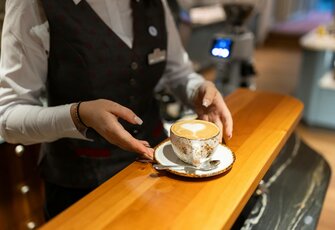 Eine Servicekraft stellt eine Tasse Cappuccino auf einem Teller auf der Holztheke ab.