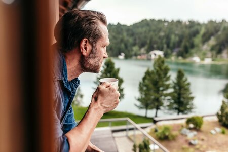 Ein Mann lehnt am Fenster und hält eine Tasse, während er auf den See und die bewaldete Uferlandschaft blickt.