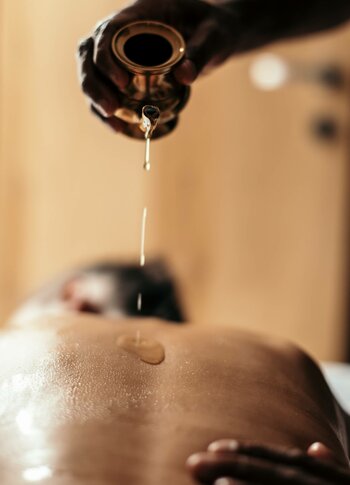 Eine Hand gießt warmes Öl auf den Rücken einer liegenden Person bei der Abhyanga-Massage mit Shirodhara.