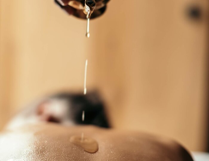 Eine Hand gießt warmes Öl auf den Rücken einer liegenden Person bei der Abhyanga-Massage mit Shirodhara.