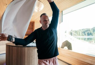 A man waves a towel in the bright panorama sauna with a view of the lake and mountains.