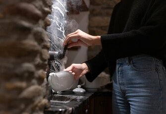 Eine Person gießt Wasser aus einem Samowar in eine weiße Teekanne an einer Tee-Station.