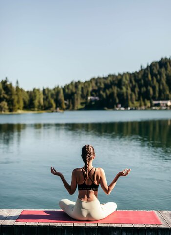 Eine Frau sitzt auf einer Yogamatte am Seeufer und meditiert mit Blick auf den Turracher See.