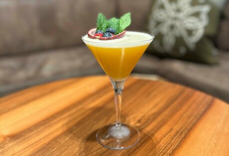 Ein gelber Cocktail mit Beeren und Minze steht auf einem Holztisch in der Hotelbar.