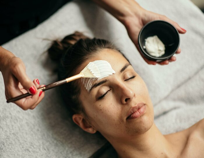 A woman receives a facial treatment while lying down, with a brush and bowl held above her face.