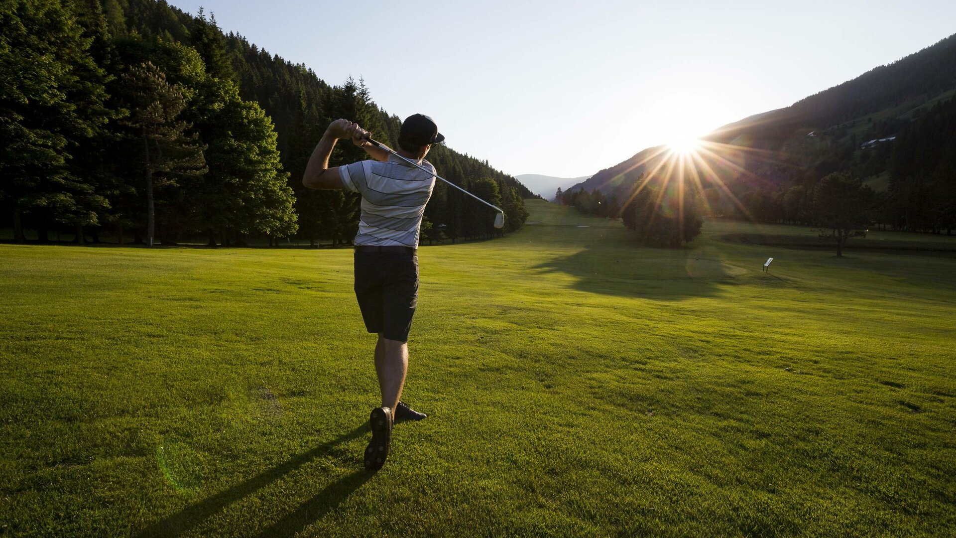 Ein Golfer schlägt auf einer weiten Wiese vor bewaldeten Hängen und tief stehender Sonne ab.