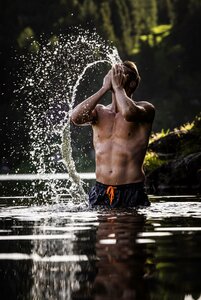 Ein Mann steht im See-Bad und splasht Wasser über sein Gesicht, während die Sonne die Tropfen beleuchtet.