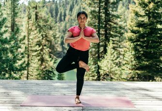 Eine Frau übt Yoga im Baum-Pose-Stil auf einer Holzterrasse vor Wald und Bergen.