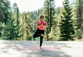 A woman practices tree pose on a wooden deck with forest and mountains in the background.