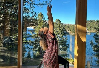 Eine Frau übt Yoga auf einer Matte vor großen Fenstern mit Blick auf den See und den Wald.