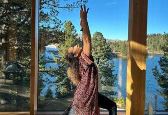 A woman practices yoga on a mat in front of large windows overlooking the lake and forest.