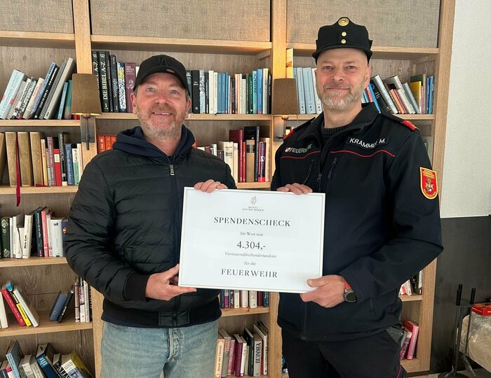 Zwei Männer halten in einer Bibliothek einen Spendencheck mit der Aufschrift „Feuerwehr“.