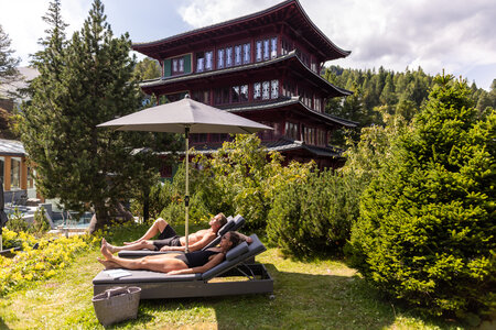 Zwei Gäste liegen auf Sonnenliegen im Garten vor dem Chinaturm, umgeben von Bäumen und Sträuchern.
