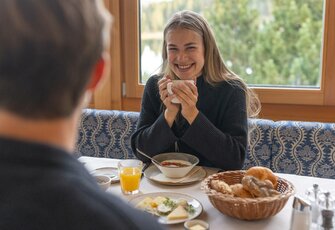 Eine Frau sitzt lächelnd am Frühstückstisch mit Suppe, Orangensaft und Brot im Hotel.