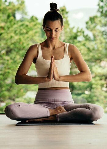 Eine Frau sitzt in der Yoga-Meditation auf einer Matte vor großen Fenstern mit Blick ins Grüne.