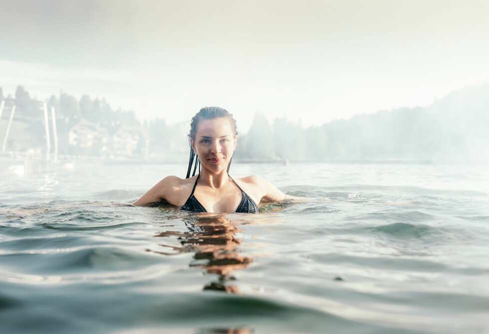A woman is swimming in the heated lake pool on Lake Turracher, with the shore and mountains visible behind her.