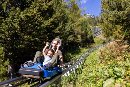 Ein Kind und ein Erwachsener fahren gemeinsam in einem blauen Sommerrodel auf einer Bergbahnstrecke durch den Wald.