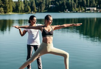 Zwei Frauen üben Yoga am Seeufer vor einer ruhigen Bergkulisse.