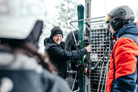 Drei Personen stehen im Schnee neben einem Wagen mit Skiern und anderen Wintersportgeräten.
