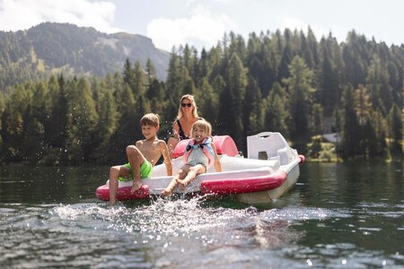 Zwei Kinder sitzen mit einer Frau auf einem Tretboot auf dem See, dahinter Wald und Berge.