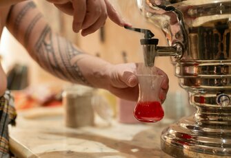 A tattooed person fills a small glass with red tea from a shiny samovar in the hamam.