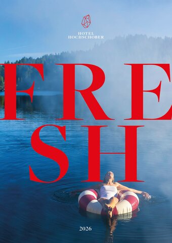 A woman lies relaxed on a red-and-white swim ring in a calm lake with wooded shores in the background.