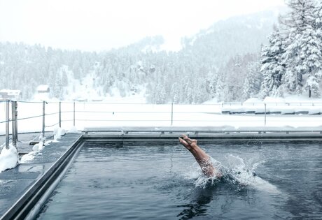 Ein Schwimmer zieht im beheizten See-Bad Bahnen, während Schnee und verschneite Berge den Hintergrund prägen.