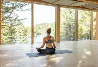 Eine Frau sitzt in meditativer Yoga-Position auf einer Matte vor großen Fenstern mit Blick auf den See.