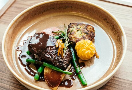 Ein angerichteter Teller mit Fleisch, Gemüse und einer Beilage liegt auf einem Holztisch im Restaurant.
