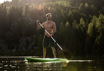 Ein Mann steht auf einem SUP-Board auf dem Turracher See vor bewaldeten Berghängen im warmen Abendlicht.