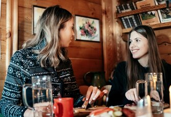 Zwei Frauen sitzen sich in einem gemütlichen Holzraum mit Getränken an einem Tisch gegenüber und lächeln einander an.