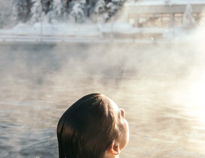 Wellness im warmen Außenbecken im Winter Eine Frau sitzt im dampfenden Außenbecken, während verschneite Bäume das winterliche Ufer im Hintergrund säumen.