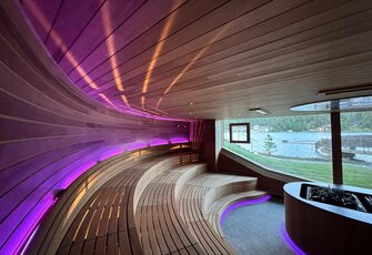 A panorama sauna with curved wooden benches, violet lighting and a view of the lake.