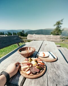Eine Hand hält einen Holzbrettteller mit Jause auf einem Holztisch mit Blick in die Berge.