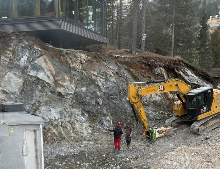 Ein gelber Bagger arbeitet an der Baustelle neben dem Hotel Hochschober am Hang.