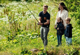 Ein Mann angelt mit einer Frau und zwei Kindern am grasbewachsenen Ufer des Turracher Sees.