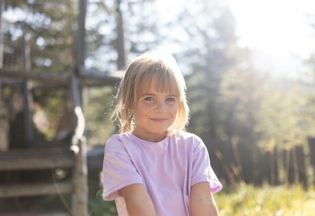 Ein Kind im rosafarbenen T-Shirt sitzt draußen in der Sonne vor einem Holzhäuschen.