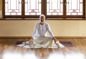 Ein Mann sitzt auf einer Yogamatte im hellen Raum vor den Fenstern und meditiert in ruhiger Haltung.
